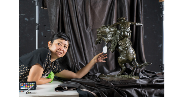 Artist Stephanie Syjuco leans on a table and holds a tag that hangs from a sculpture.