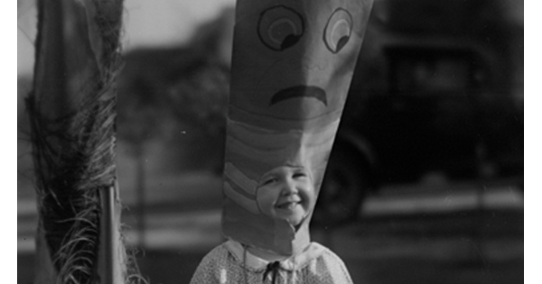 A black-and-white photo of a child wearing a tall, cardboard hat with a face painted on the top.