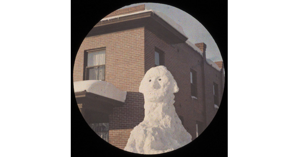 A color photograph of a cartoonish likeness of George Washington sculpted from snow in front of a red brick building. 