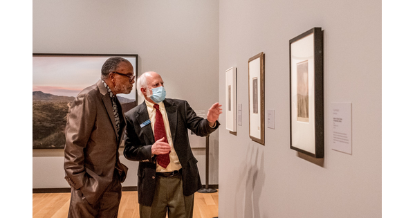 Two men stand in a gallery looking at artwork; one man gestures toward the art