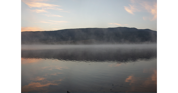 A detail of a color photograph of morning fog over a body of water with dark hills in the background.