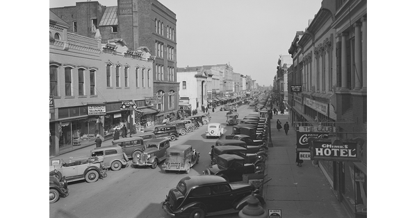 A black-and-white image of a street lined with tall buildings and parked automobiles.