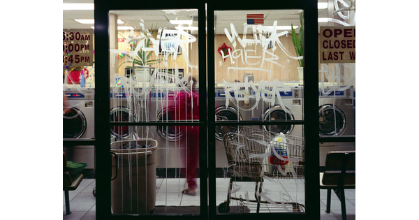 A color photograph of a laundromat at night as seen through glass doors covered in white graffiti.