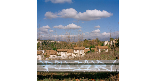 A color photograph of a graffitied guardrail with houses in the background under a cloud-filled sky.