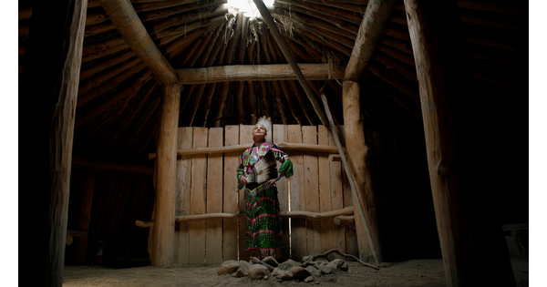 A screenshot of an Indigenous woman in traditional dress standing in a wood structure looking up at an opening to the sky.