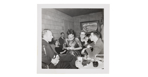A black-and-white snapshot of formally-dressed people at a party in a cinderblock room.