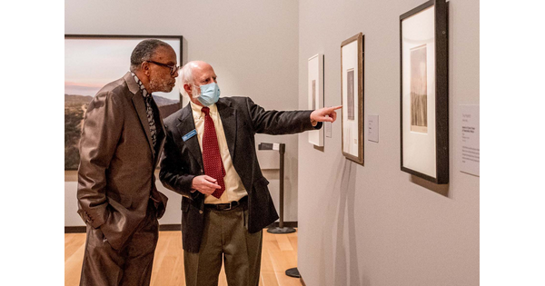 Two men, one pointing at a photo on the wall, stand in a gallery of framed photographs.