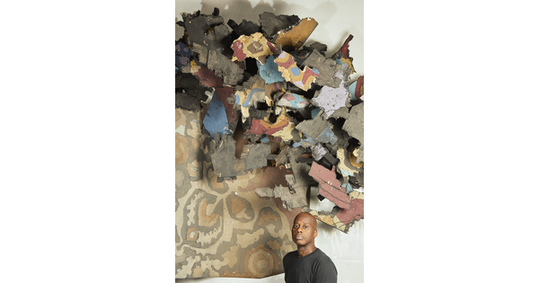 A Black man stands under an abstract installation high on a wall.