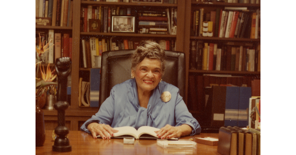 A color photo of an older woman sitting at a desk in a room lined with floor-to-ceiling bookshelves.