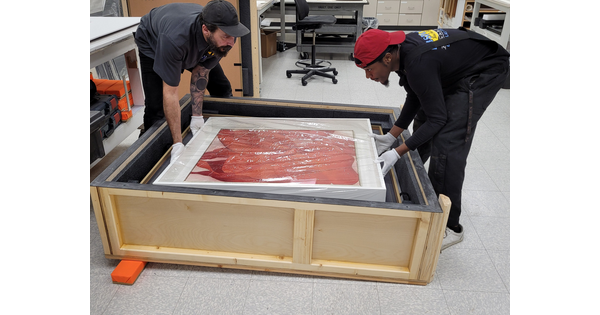 Two people carefully place a painting inside of a wooden crate.