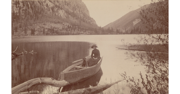 A sepia-toned photograph of a woman and her dog sitting in a canoe on a very still lake set in the mountains.