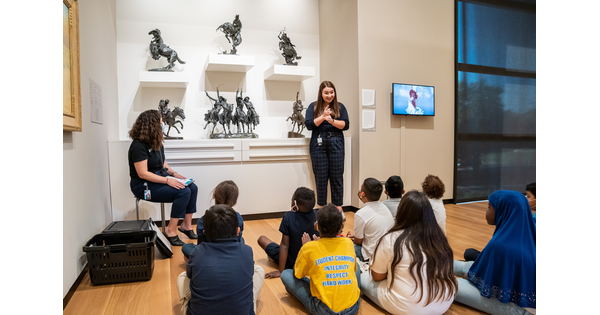 A school group sits on the floor in a Carter gallery watching an ASL interpreter sign during a tour.