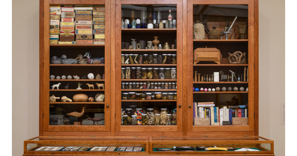 A detail of a large wooden cabinet with drawers on the bottom half and glass-fronted cabinets above, all filled with random objects.