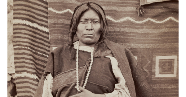 A sepia-toned photograph of an Indigenous person seated in front of woven blankets with their thumb hooked over a white beaded necklace hanging from their neck.