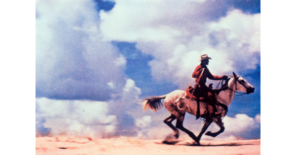 A color photograph of a man on a running horse in a dusty landscape and a sky full of white, fluffy clouds.