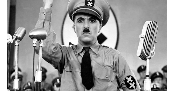 A black-and-white film still showing a White man dresses as Adolf Hitler standing at a microphone and gesturing with his arms.