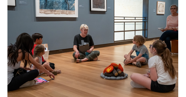 Children and adults sit on the floor of a Carter gallery around a pretend campfire listening to an educator.