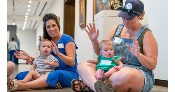 Two adult women sit cross-legged on the floor of a Carter gallery, each with a baby on their lap.