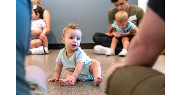 A baby sits on the floor of a Carter gallery surrounded by other adults with babies.