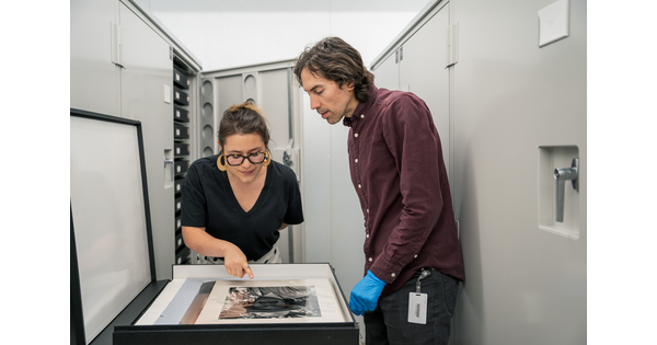 A light-skinned woman leans over a box of photographs in a storage space as a light-skinned man watches.