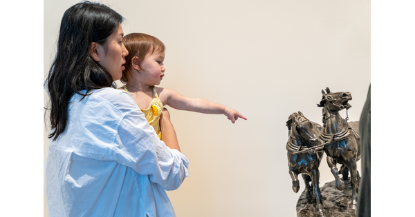 Woman holds toddler pointing at bronze sculpture of rearing horses.