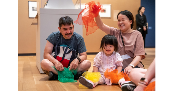 Two adults and a child sit on the floor of a Carter gallery playing with and waving colorful fabric.