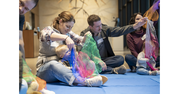 Adults and babies sit on the floor of a Carter gallery playing with colorful pieces of sheer fabrics.
