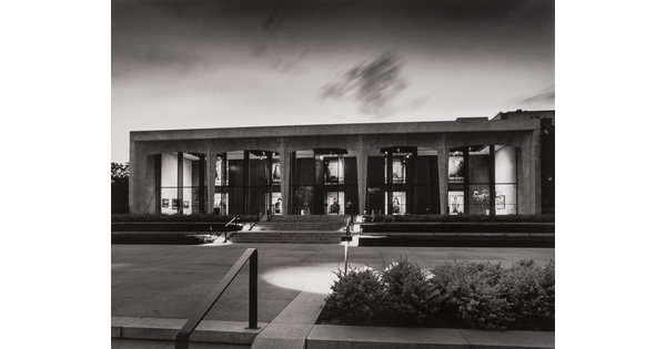 A black-and-white photograph of the facade of the Carter at dusk; large-scale portrait photographs can be seen through the Carter&#039;s windows.