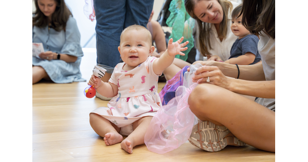 A White baby sits on the floor of a Carter gallery, adults nearby, waving and smiling.