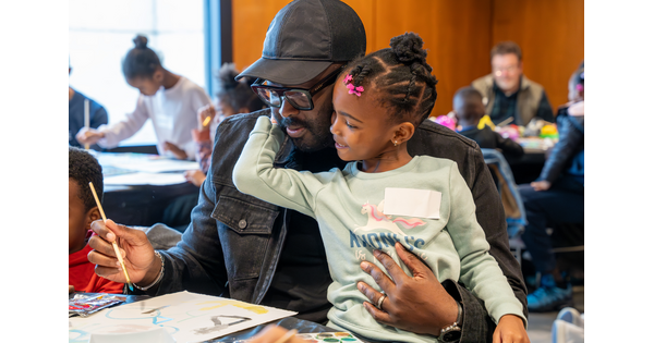 A smiling Black child sits on the lap of a Black man as they paint together in the Carter&#039;s Pop-up Lounge.