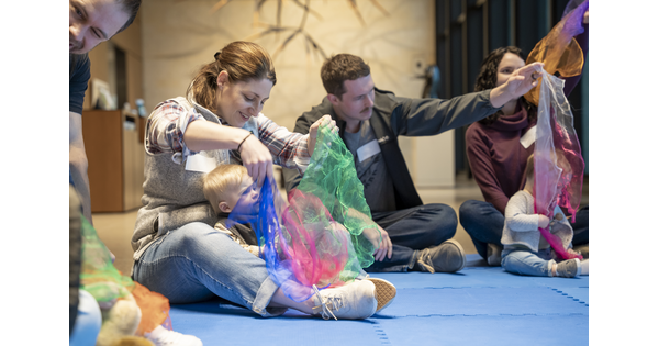 Several adults sit on the floor of a Carter gallery with their babies in their laps playing with colored cloths.