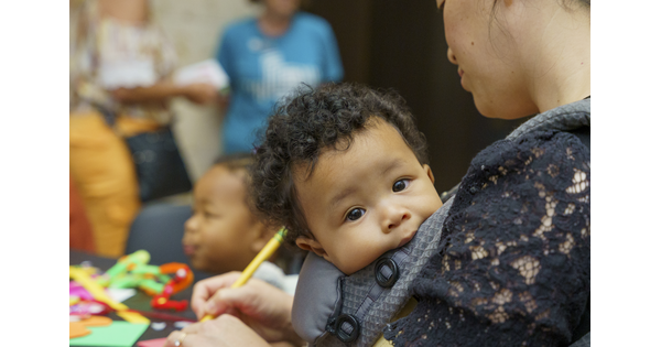 A baby with a full head of dark curly hair rests in a sling as the adult holding it makes art.