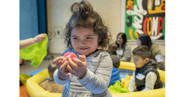 A White child with dark curly hair holds a toy in her hands as she plays in a Carter gallery.