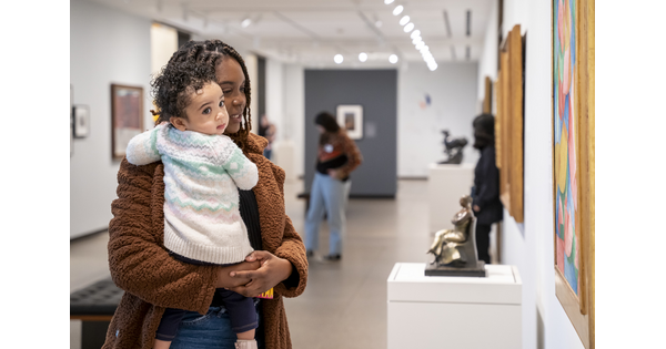 A Black woman holding a baby in her arms looks at a sculpture in a Carter gallery.