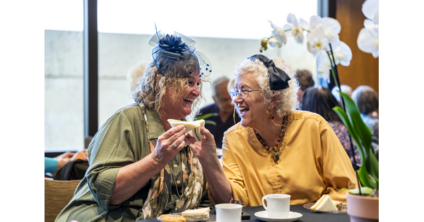 Two older White women, both wearing hats, laugh together as they enjoy sandwiches.