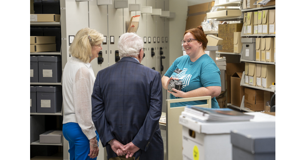 A smiling young White woman with red hair shows an object to a couple standing in a room lined with shelves..