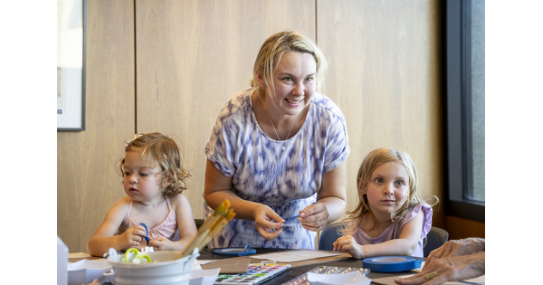 A White woman leans over a table in the Carter's Lounge where two children are making artwork.