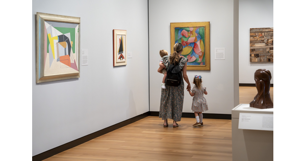 A woman and two children, one she holds on her hip and she hold the hand of the other, look at colorful art in a Carter gallery.