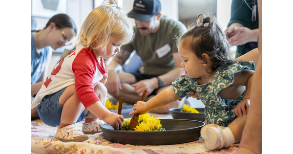 Two very young children play together with colored flowers in a bowl on the floor of a Carter gallery.