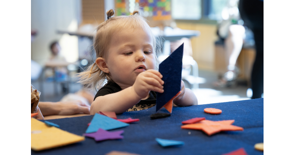A small White child at a table playing with colored felt in the shapes of stars and triangles.
