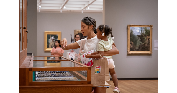 A dark-skinned woman holding a child on her hip points to some of the objects in "The Texas Cabinet."
