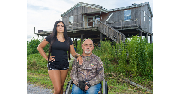 Two people outside of a house on stilts: a young woman with long dark hair stands next to a middle-aged man seated in a wheelchair.