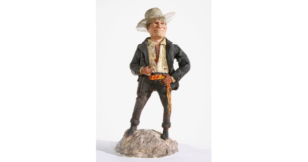 A painted sculpture of a White man wearing a cowboy hat, a light-colored shirt, and dusty pants and jacket standing on a rock, smoking a cigarette.