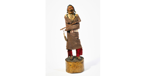 A painted wood sculpture of a smiling Native American man with exaggerated features wrapped in a blanket with a pipe resting on his arm.