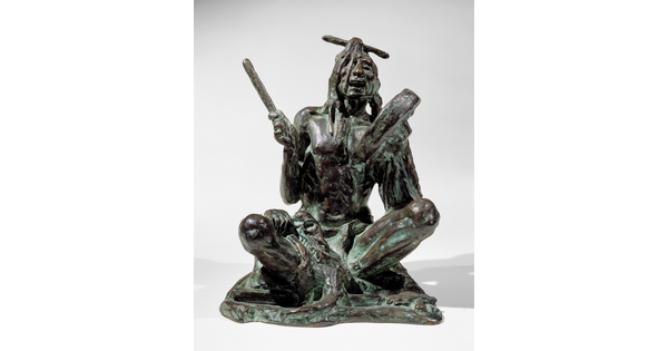 A bronze sculpture of a sitting Indigenous man playing a handheld drum.