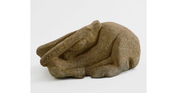 A stone sculpture of a goat curled on the ground with its head turned so its chin rests on its shoulder.