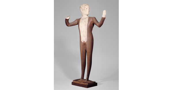 A wood sculpture of a male orchestra conductor standing with his arms raised out to his side.