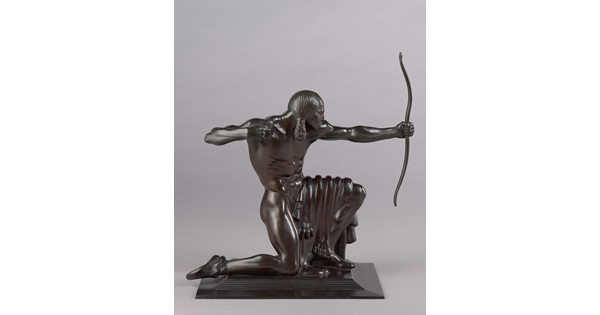 A bronze sculpture of a stylized Native American man on one knee pulling back the string of a bow he holds in front of him.