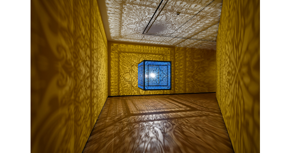 A brightly lit and suspended blue cube with laser cut shapes on all sides that cast intricate shadows on yellow walls.