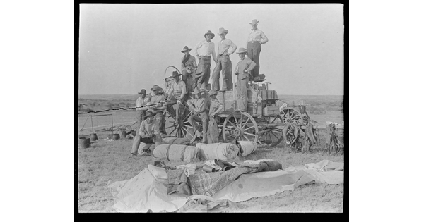 A black-and-white image of a group of cowboys posed on a wagon, bedrolls and blankets on the ground in front of them.